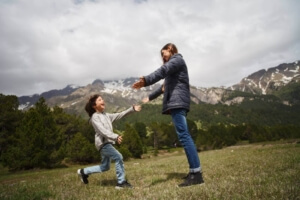 Happy Woman and Child Running in a Scenic Field with Majestic Mountains in the Background